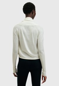 Suéter de punto color crema con textura suave, que presenta un cuello tipo chal y puños acanalados. Incluye un detalle metálico cerca del dobladillo.