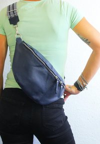 Sac en bandoulière en cuir bleu avec une finition texturée et des fermetures à glissière argentées ; la sangle à motifs réglable apporte une touche de détail. Porté sur la hanche contre une chemise vert clair.