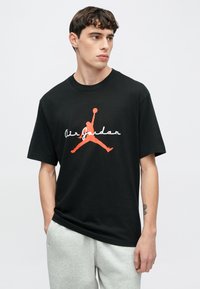 Μαύρο βαμβακερό t-shirt με κόκκινο λογότυπο Jumpman και τη φράση "Air Jordan" σε λευκό στυλ γραφής. Χαλαρή εφαρμογή, στρογγυλή λαιμόκοψη, φορεμένο με ανοιχτό γκρι φόρμα.