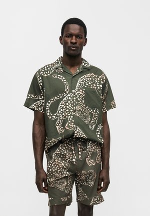 Άνδρας που φοράει ταιριαστό ελιάς κοντομάνικο πουκάμισο και σορτς με μπεζ πουα λεοπάρ μοτίβο, στέκεται μπροστά σε απλό φόντο.