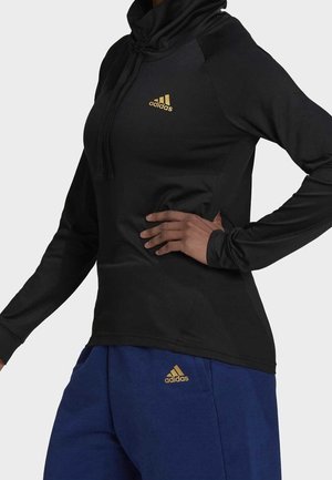 Person trägt schwarzen Adidas Hoodie mit langen Ärmeln und blaue Adidas Hose mit goldenen Logos, Hand ruht auf der Hüfte.