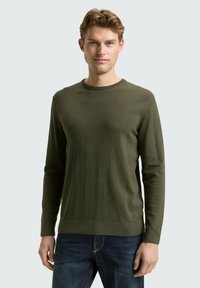 Groene gebreide trui met een ronde hals en lange mouwen, met een gladde textuur en een aansluitend ontwerp. Draagt over blauwe spijkerbroek.