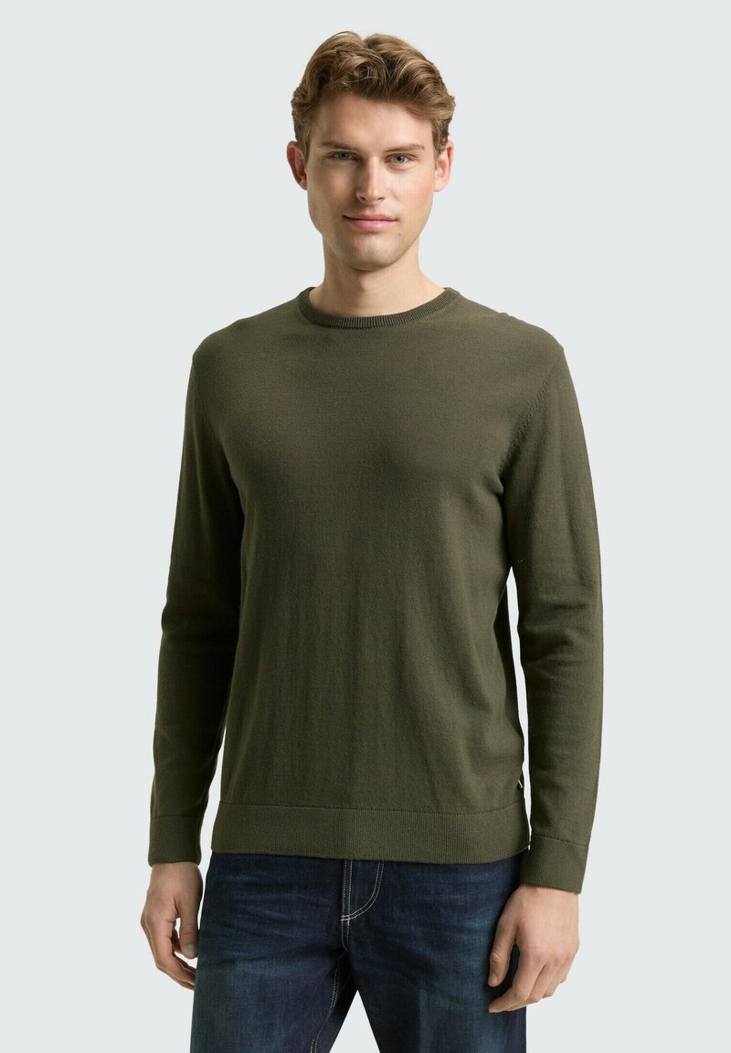 Groene gebreide trui met een ronde hals en lange mouwen, met een gladde textuur en een aansluitend ontwerp. Draagt over blauwe spijkerbroek.