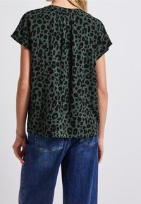 Top verde a stampa leopardata con maniche corte e dettagli plissettati sul retro, abbinato a jeans denim blu. Il tessuto appare liscio e leggero.