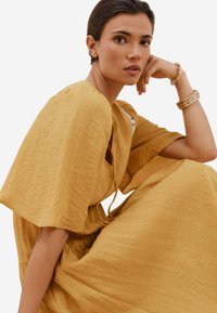 Senfgelb, strukturiertes Leinenkleid mit weiten Ärmeln, Kordelzug-Ausschnitt und Seitentaschen. Modell trägt ein mehrsträngiges Armband.
