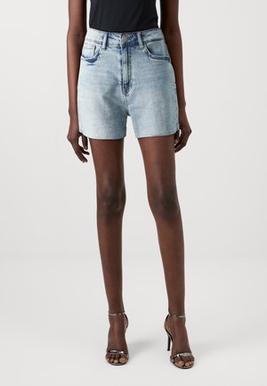 Femme portant un short en jean bleu clair, un top noir sans manches et des sandales noires à talons hauts avec brides, debout devant un fond uni.