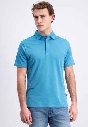 Jonge man met krullend bruin haar, gekleed in een lichtblauw polohemd met korte mouwen en blauwe jeans, staand tegen een simpele witte achtergrond.