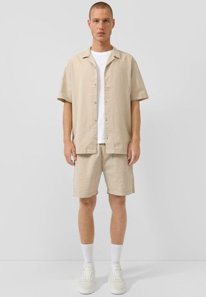 Mann trägt beiges kurzärmliges Hemd mit Knöpfen und passende Shorts, weißes T-Shirt darunter, weiße Socken und weiße Sneakers, steht vor einer grauen Wand.