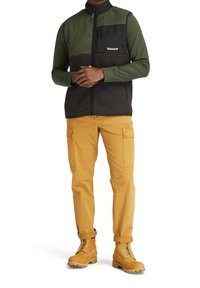 Fleeceväst i mörkgrön och svart, dragkedja fram, en bröstficka. Kombinerad med senapsgula cargobyxor och tanfärgade kängor med snörning.