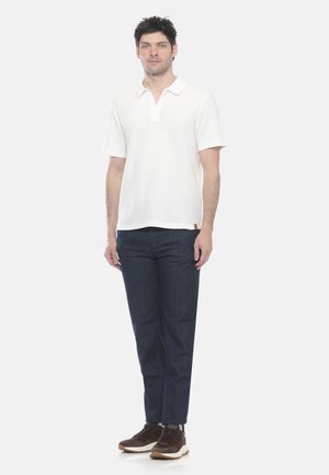 Uomo in piedi che indossa una polo bianca a maniche corte, jeans blu scuro e sneakers marroni su uno sfondo chiaro uniforme.