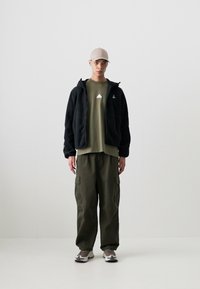 Casaco de fleece preto com fecho éclair, t-shirt verde-oliva com um pequeno gráfico, calças cargo e ténis em tons neutros. Conjunto simples e funcional.