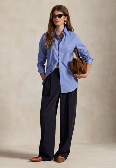 Γυναίκα που φοράει γυαλιά ηλίου από καβούκι χελώνας, μπλε πουκάμισο με κουμπιά, χαλαρούς μαύρους παντελόνια, καφέ μπαλαρίνες και κρατάει μια καφέ δερμάτινη τσάντα.