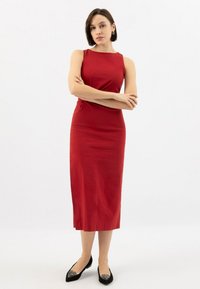 Donna dai capelli corti che indossa un vestito midi rosso senza maniche e delle ballerine nere con decorazioni in strass, in piedi con le braccia incrociate su uno sfondo bianco.