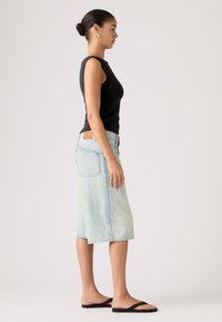 Γυναίκα που στέκεται πλάγια, φοράει μαύρο αμάνικο τοπ, φούστα από denim μήκους μέχρι το γόνατο σε ανοιχτό μπλε χρώμα, μαύρα σαγιονάρια και χρυσούς κρίκους σκουλαρίκια.