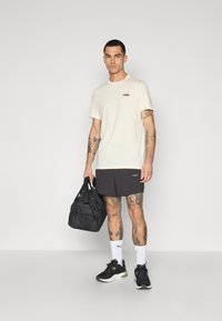 Lys beige t-skjorte med korte ermer og rødt logo, kombinert med svarte shorts. Mørke joggesko, hvite sokker og en svart treningsbag. Tatoveringer synlige.