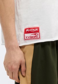 Λευκό T-shirt με κόκκινο σήμα "VENUM TYSON" στο τελείωμα; συνδυάζεται με πράσινο και καφέ αθλητικό παντελόνι με πλαϊνές λεπτομέρειες. Μαλακό ύφασμα τζέρσεϊ.