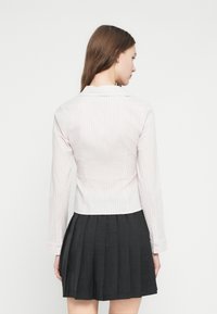 Γυναίκα με μακριά καστανά μαλλιά που φοράει ανοιχτόχρωμο ριγέ πουκάμισο με μακριά μανίκια και μαύρη πλισέ φούστα, στέκεται με την πλάτη προς το μέρος μας μπροστά σε λευκό φόντο.