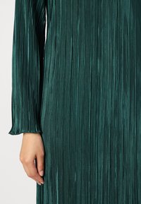 Τιρκουάζ φόρεμα με πτυχώσεις και μακριά μανίκια, που διαθέτει κάθετους ρίγες και λεία υφή, αναδεικνύοντας μια εφαρμοστή σιλουέτα και ρέουσα γραμμή.