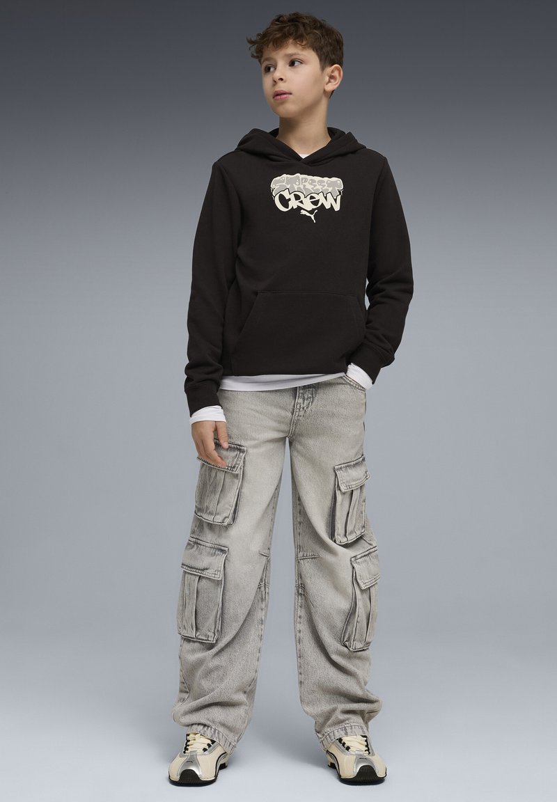 Garçon portant un sweat à capuche noir avec le logo "Crew", un pantalon cargo gris clair et des baskets couleur crème, debout devant un fond dégradé gris.