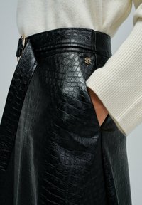 Jupe en simili cuir noir avec motif en relief de crocodile, ceinture avec boucle dorée, et main dans la poche, associée à une manche de pull côtelé crème.