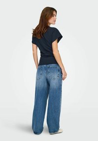 Femme aux longs cheveux bruns portant un haut noir à manches courtes et un jean bleu à jambes larges, debout et regardant sur le côté.