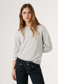 Maglione grigio chiaro con scollatura rotonda e polsini a coste, abbinato a jeans scuri con chiusura a due bottoni e dettagli di cucitura sottili.