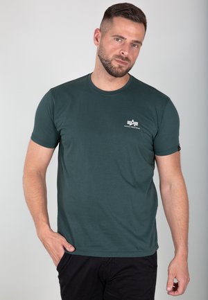 Camiseta verde oscuro de algodón, con cuello redondo, mangas cortas y un pequeño logo blanco en el lado izquierdo del pecho. Textura suave.