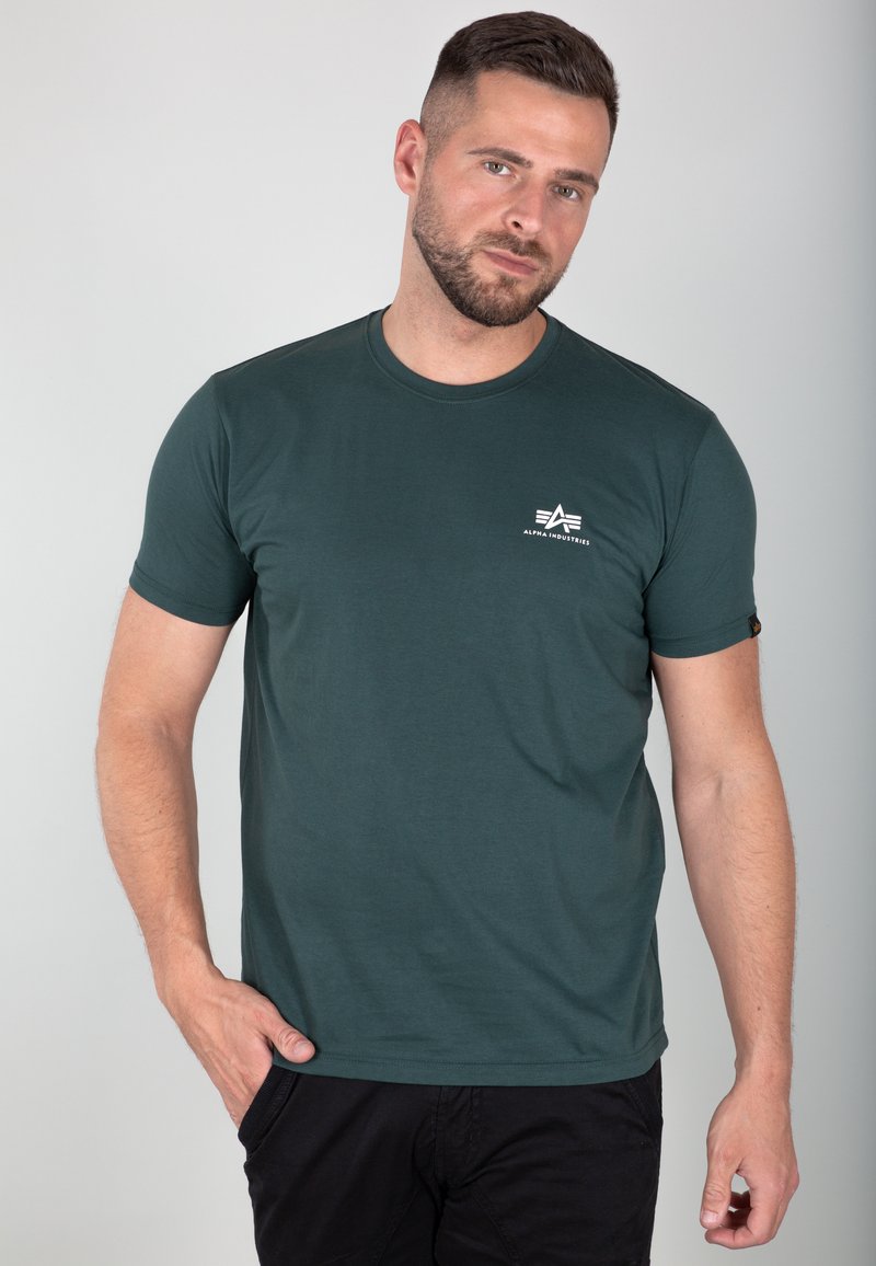 Dunkelgrünes T-Shirt aus Baumwolle, mit Rundhalsausschnitt, kurzen Ärmeln und einem kleinen weißen Logo auf der linken Brust. Glatte Textur.