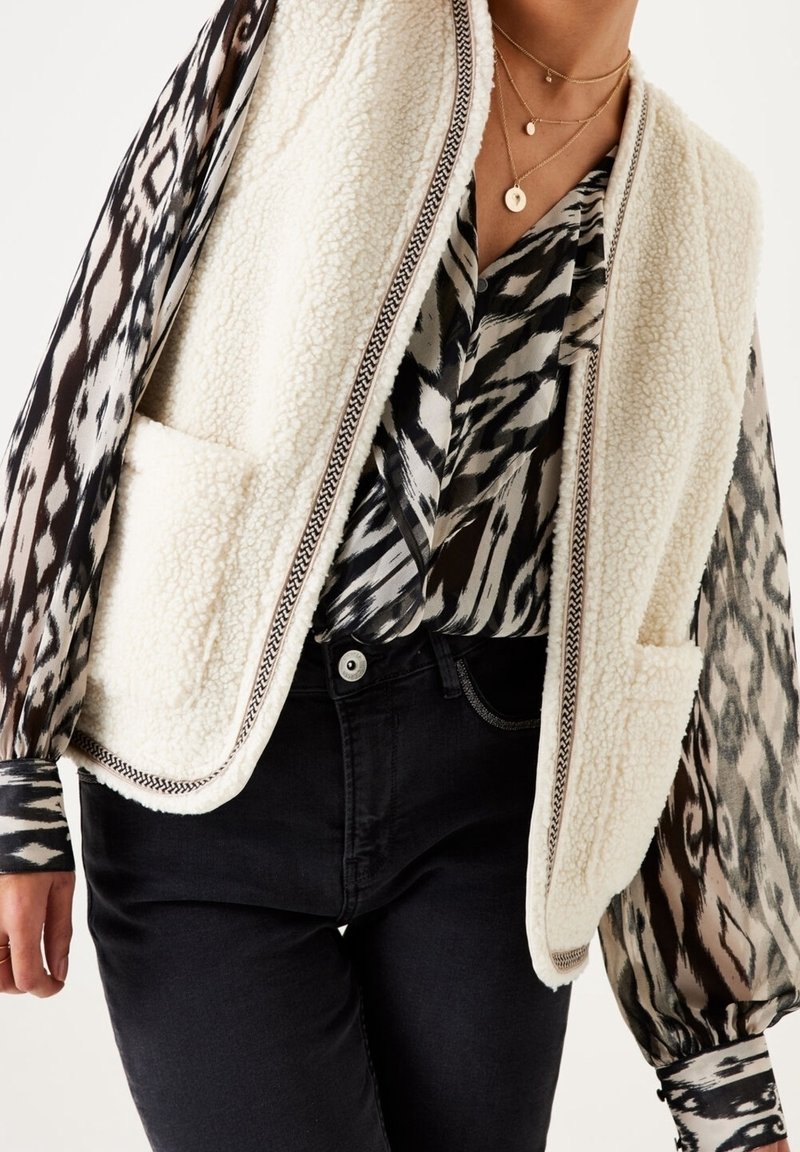 Fleeceväst i krämfärgad med en strukturerad yta, öppen front och två fickor. Kombineras med en mönstrad svart och vit blus och svarta jeans.