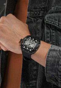 Zwarte, vierkante horloge met een skeleton wijzerplaat en rubberen band; bevat duidelijke details van de interne mechanismen en zilveren accenten.