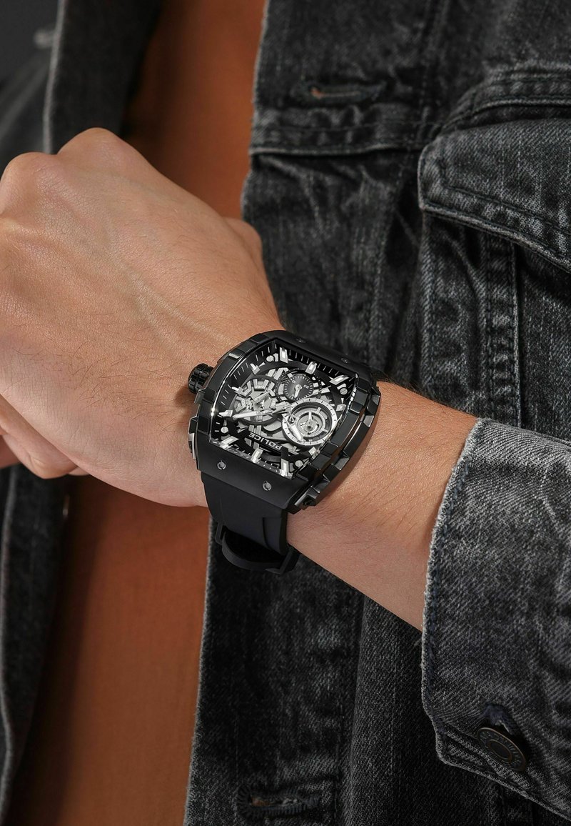 Zwarte, vierkante horloge met een skeleton wijzerplaat en rubberen band; bevat duidelijke details van de interne mechanismen en zilveren accenten.