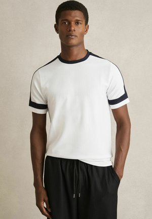 Tricou alb cu mânecă scurtă, cu dungi, având garnituri de contrast bleumarin la guler și mâneci, purtat cu pantaloni negri cu șnur.
