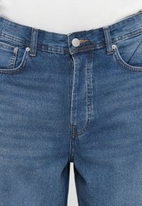 Krupan kadar plavih traperica na struku s metalnim gumbom, patentnim zatvaračem, petljama za pojas i prednjim dzepovima, koje su kombinirane s bijelom majicom.