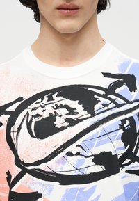 Κοντινό πλάνο ενός ατόμου που φοράει λευκό πουκάμισο με ένα μεγάλο μαύρο σήμα της Nike που καλύπτει μια σχεδίαση υδρογείου σε μαύρο, μπλε και πορτοκαλί.