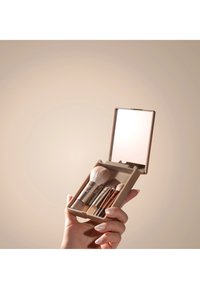 Hånd som holder et kompakt sett med sminkekoster med fire koster og et åpent speil mot en beige bakgrunn.