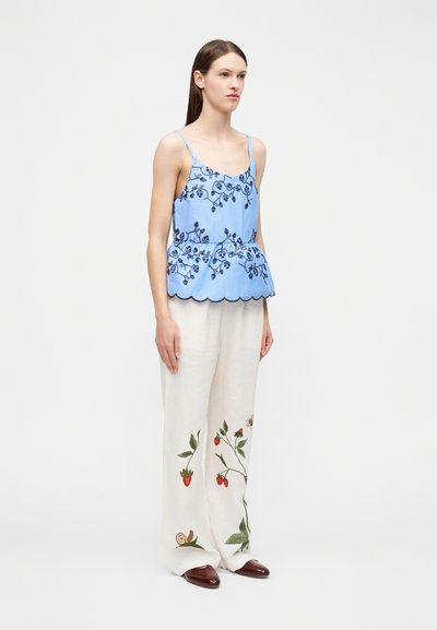 Γυναίκα που φοράει μπλε αμάνικο τοπ με λουλουδάτο σχέδιο και λευκό παντελόνι φαρδιάς γραμμής με στάμπες φράουλας, μέλισσας και σαλιγκαριού, στέκεται μπροστά σε μονόχρωμο φόντο.