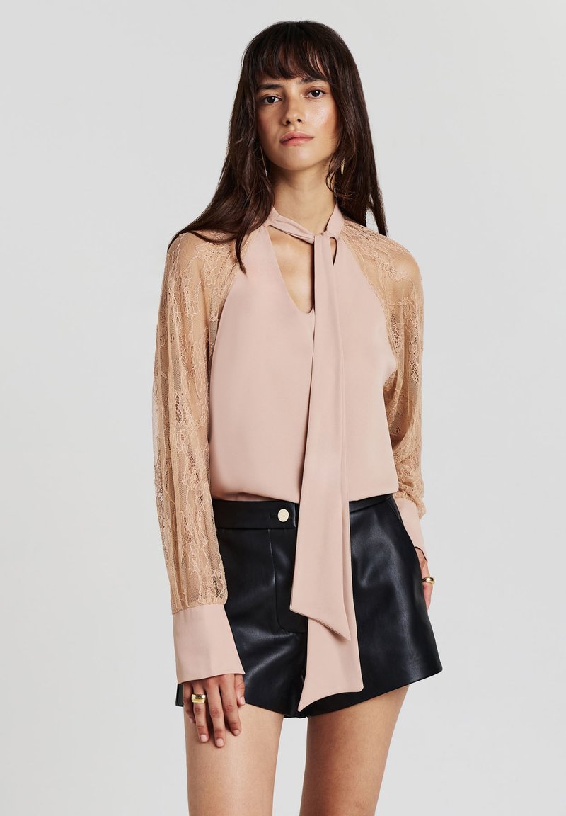 Frau, die eine blush-pinke Bluse mit Spitzenärmeln und Halsbanddetail trägt, kombiniert mit schwarzen Leder-Shorts, vor einem schlichten Hintergrund stehend.