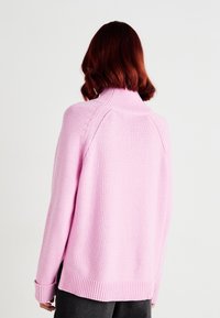 Pull rose clair à col montant, poignets et ourlet côtelés. Il présente un tricot texturé et des coutures d'épaule tombantes pour une coupe décontractée.