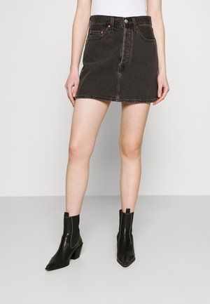 Femme portant une mini-jupe en jean noire, un haut blanc et des bottines noires, debout devant un fond clair uni.