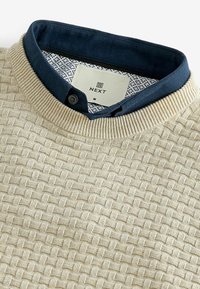 Pull en tricot beige avec un tissage texturé, doté d'un col foncé bleu et d'un doublure intérieure à motifs. Étiquette de la marque visible.