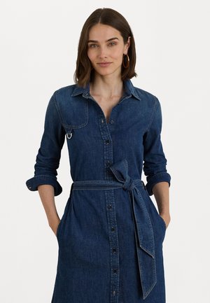 Kvinne med skulderlangt brunt hår iført en belted langermet denimkjole med oppbrettede ermer og gull hoopsøreringer, hendene i lommene.