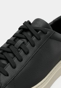 Sorte lær sneakers med teksturert overflate, flate sorte snøring og en kremfarget gummisåle. Har preget logo på siden.