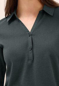 Tmavě zelená blůza s dlouhým rukávem, límečkem a třemi knoflíky. Měkká tkanina s hladkým povrchem a volným střihem.