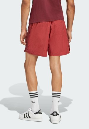 Male wearing red athletic shorts, maroon shirt, white Adidas socks, and white Adidas sneakers standing facing away on a plain background.