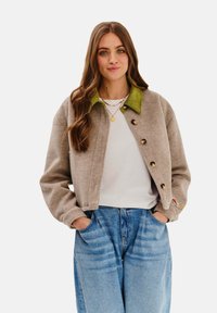 Krótkiażdżowa kurtka w kolorze tan z zielonym kołnierzem z kordurowego materiału, wyposażona w duże guziki i luźny krój. Zestawiona z niebieskimi dżinsami o wysokim stanie.