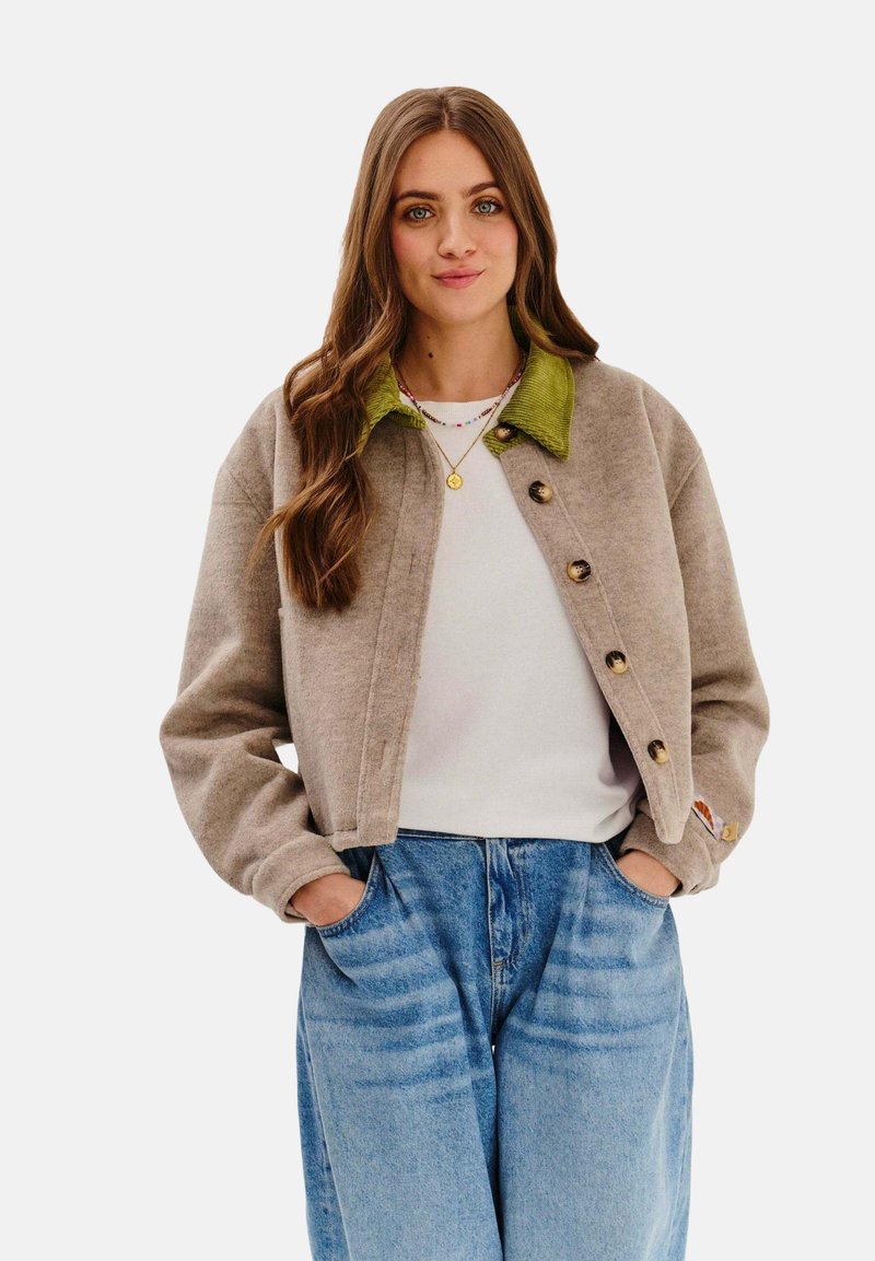 Krótkiażdżowa kurtka w kolorze tan z zielonym kołnierzem z kordurowego materiału, wyposażona w duże guziki i luźny krój. Zestawiona z niebieskimi dżinsami o wysokim stanie.