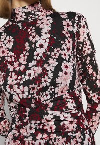 Pitkähihaista puseroa mustasta kankaasta, jossa on kukkakuosi vaaleanpunaisena ja punaisena. Korkea kaulus ja kootut yksityiskohdat vyötäröllä.