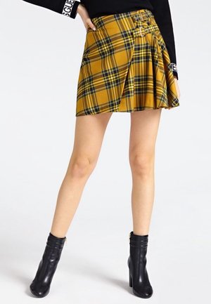 Woman wearing a yellow plaid pleated mini skirt with buckle details, black sweater, and black ankle boots standing with legs crossed.