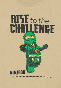 Grüner Ninjago-Charakter auf einem beigefarbenen T-Shirt mit dem Text "RISE to the CHALLENGE" in fetter schwarzer Schrift, das detaillierte Designelemente aufweist.