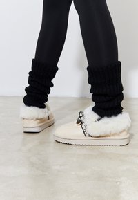 Pantofole beige con bordatura in pelliccia bianca, dettaglio della fibbia nera e scaldamuscoli neri testurizzati. Materiale morbido e suola piatta. Superficie liscia.